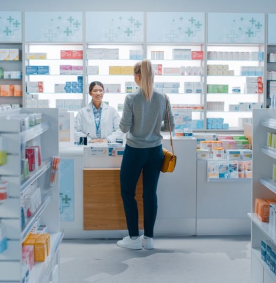 Pharmacy Drugstore: Beautiful Young Woman Buying Medicine, Drugs, Vitamins Stands next to Checkout Counter. Female Cashier in White Coat Serves Customer. Shelves with Health Care Products